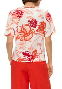 Bloemige blouse in het wit, met rode en roze bloemenpatronen, korte mouwen, ronde halslijn, en een ontspannen pasvorm, gecombineerd met rode broeken.