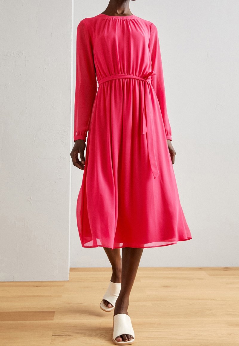 Femme marchant à l'intérieur, vêtue d'une robe midi à manches longues rose vif avec une ceinture à la taille et des sandales mules ouvertes blanches sur un sol en bois.