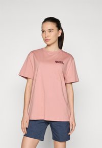T-shirt de algodão rosa com mangas curtas e decote redondo, apresentando um logótipo escuro na área do peito esquerdo. Combinado com calções azuis marinhos.