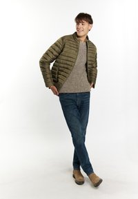 Giacca trapuntata verde oliva con colletto alto, indossata sopra un maglione di maglia beige. Jeans blu e stivali in camoscio marroni completano l'outfit.