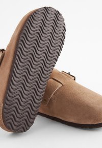 Brūnas zamša klumpes ar teksturētu tumšu gumijas zoli, ar metāla sprādzes detaļu un gludu, apaļu pirkstu dizainu.