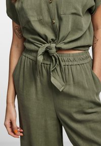 Zelený lněný dvoudílný set obsahující uzlový top s krátkým rukávem a knoflíky a kalhoty s elastickým pasem a kapsami. Hladká textura.