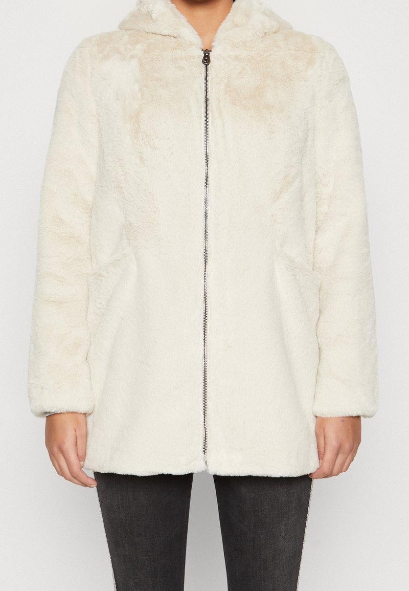 Personne portant une veste crème duveteuse à fermeture éclair, manches longues et col légèrement relevé, associée à un pantalon foncé.