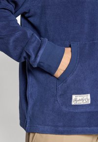 Maglione blu navy con tessuto testurizzato, tasca a marsupio e polsini a coste. Presenta un'etichetta decorata nell'angolo inferiore.
