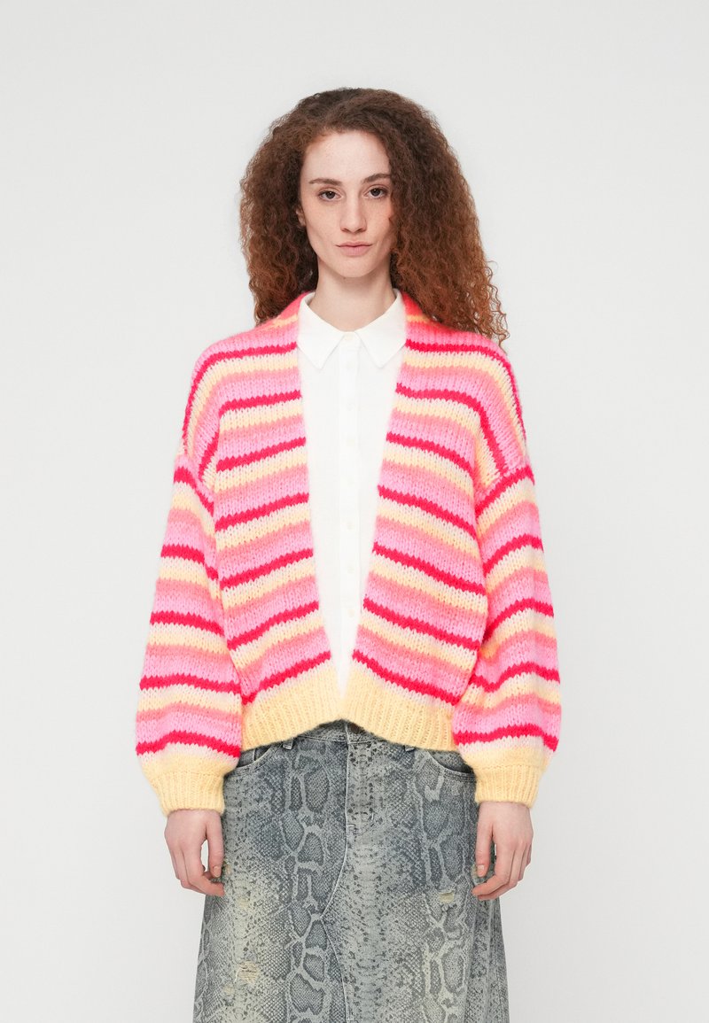 Πολύχρωμη ζακέτα με φαρδιές ροζ, κόκκινες και κίτρινες ρίγες, πλεκτή υφή, ανοιχτό μπροστινό μέρος και φουσκωτά μανίκια. Συνδυασμένη με λευκό πουκάμισο.