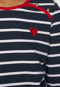 Marinblå och vit randig tröja med röd halsringning, guldknapp-dekorationer och ett rött broderat hjärta på vänster sida.