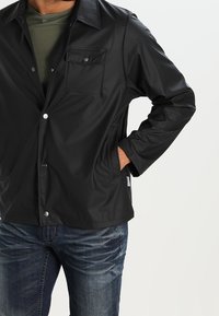 Svart vattentät jacka med en slät yta, tryckknappar och en bröstficka; bärs över en grön skjorta och blå jeans.