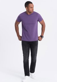 T-shirt in cotone viola con collo a giro abbinato a jeans scuri e sneakers bianche. Il modello è in piedi in modo casuale con le mani sui fianchi, mostrando il suo fit.