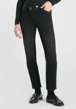 Schwarze, schmal geschnittene Jeans aus Denim mit hoher Taille, zwei vorderen Taschen und Metallknopfverschluss; kombiniert mit schwarzen Slippern und Socken.