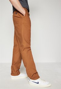 Brown cotton trousers with a straight fit, featuring side pockets and a small logo patch on the waistband. Paired with white sneakers.