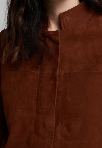 Bruine suède top met een hoge, ronde kraag, voorzien van verticale stiksels en een gladde textuur. Geen zichtbare sluitingen of versieringen.