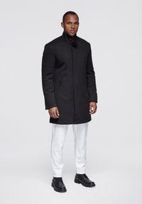 Zwarte jas met een hoge kraag, zijzakken met rits en een rechte snit, gecombineerd met witte broek en zwarte schoenen. Glad oppervlak.