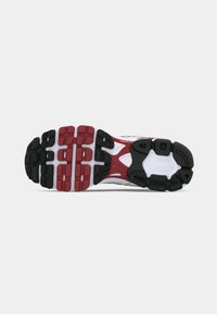 Suela exterior de zapato deportivo con goma negra y detalles en rojo y blanco; patrón texturizado con ranuras profundas para tracción y flexibilidad.