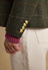 Close-up van een geruite groene jas mouw met vier gouden knopen boven een roze trui manchet en een zichtbare witte broekzak.