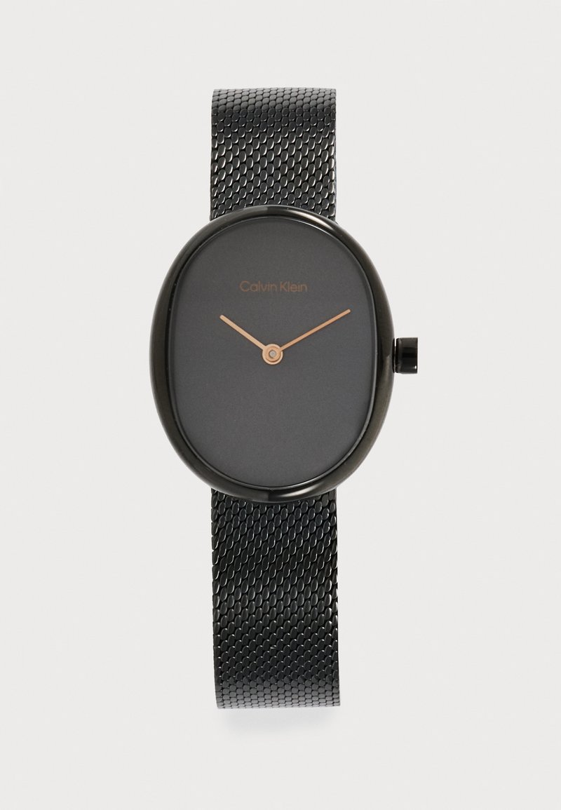 Montre ovale noire Calvin Klein avec bracelet en maille noire et aiguilles horaires et minutes dorées minimalistes sur un cadran sombre.
