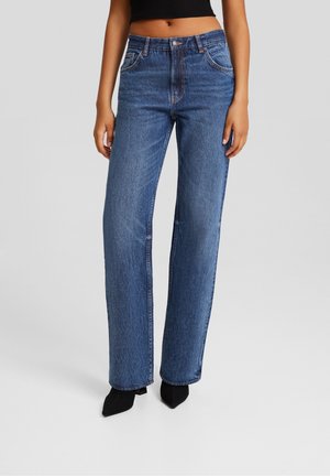 Femme vêtue d'un jean bleu droit taille haute, d'un top noir court et de talons noirs à bout pointu, debout devant un fond gris uni.