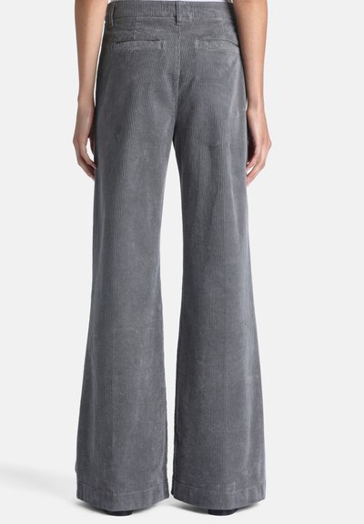 Personne portant un pantalon gris en velours côtelé taille haute à jambes larges avec poches passepoilées à l'arrière, debout devant un fond clair uni.