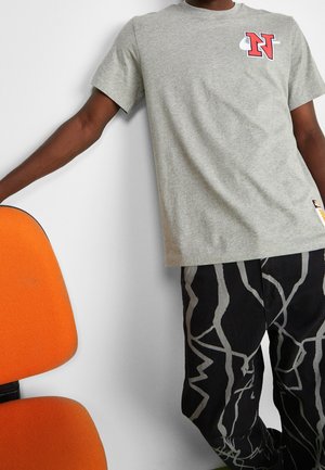 Osoba ubrana w szarą koszulkę z czerwonym logo "N" i czarne spodnie z abstrakcyjnymi białymi liniami, opierająca się o pomarańczowe krzesło.