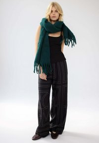 Schwarze weit geschnittene Hose mit weißen geometrischen Mustern, kombiniert mit einem schwarzen ärmellosen Oberteil und einem großen grünen Schal. Der Schal hat fransige Kanten.
