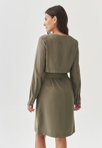 Olive green dress with long sleeves and a fitted waist, featuring a cropped jacket style and button accents on the sleeves. Smooth texture.