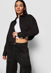 Black cropped button-up shirt with long sleeves, paired with dark cargo pants. The shirt features a classic collar and is made of smooth fabric.