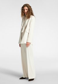 Mujer con cabello rizado vistiendo un traje de pantalón color blanco roto y zapatos marrones de punta, de pie y mirando hacia la cámara sobre un fondo claro y liso.