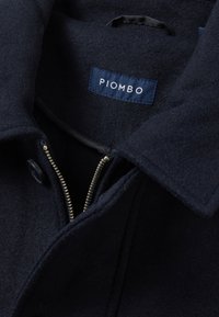 Cappotto in lana blu navy con colletto a revers, dotato di chiusura con zip e bottone, e un'etichetta in tessuto con la scritta "PIOMBO" ricamata in bianco.
