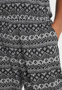Gros plan sur la main d’une personne dans la poche d’un pantalon à motifs noir et blanc présentant des formes géométriques et des dessins linéaires complexes.