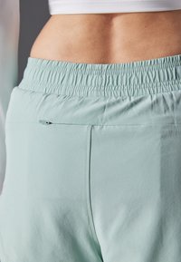 Heledad rohelised spordipüksid elastse talje ja väikese lukuga taskuga seljal, näidates sujuvat tekstuuri ja saavutades vormikohaseist fit'i.