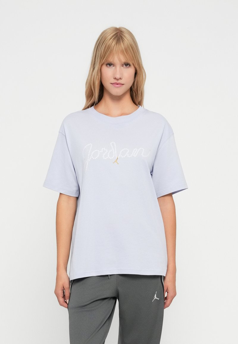 Jonge vrouw met blond haar, gekleed in een lichtpaarse Jordan T-shirt en grijze Jordan sweatpants tegen een witte achtergrond.