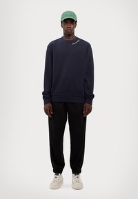 Granatowy sweatshirt z logo na piersi, czarne dresy z prążkowanymi mankietami, białe sneakersy i zielona czapka. Materiał z mieszanki bawełny, luźny krój.