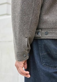 Braune Wolljacke mit strukturiertem Oberflächen, ausgestattet mit metallischen Druckknöpfen an den Ärmeln und an der Seite, kombiniert mit dunklen Jeans.