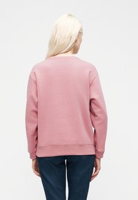 Γυναίκα με μακριά ξαν θά μαλλιά που φοράει ένα απλό ροζ φούτερ και σκούρο μπλε τζιν, εμφανιζόμενη από πίσω σε λευκό φόντο.