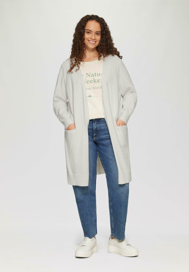 Hellgrauer Cardigan mit gerippten Bündchen und Saum, der über einem cremefarbenen T-Shirt und blauen Jeans getragen wird, kombiniert mit weißen Sneakern.