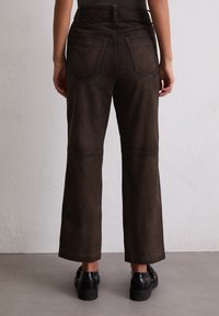 Braune Wildleder-Hosen mit weitem Bein, die über Gesäßtaschen, Nähte entlang der Beine und eine hohe Taille verfügen, kombiniert mit schwarzen Schuhen.