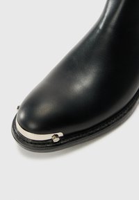 Bota de cuero negra con punta redondeada, con acentos de hardware plateado y una punta blanca en contraste. Textura suave con detalles de costura.