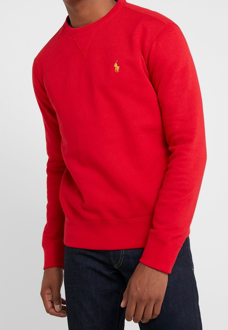 Homme portant un sweat-shirt à col rond rouge vif avec un petit logo cheval jaune sur la poitrine, associé à un jean bleu foncé.