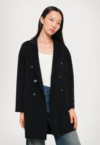 Schwarzer doppelt brustiger Mantel mit Reverskragen, zwei vorderen Taschen und glattem Textil, kombiniert mit einem weißen Oberteil und Jeans.