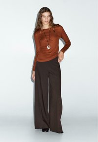 Brown long-sleeve top with a fitted design, paired with loose, wide-leg dark trousers. The outfit is complemented by a silver pendant necklace.