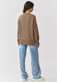 Brązowy sweter z dzianiny z prążkowanym dołem i mankietami, opadającymi ramionami oraz bocznymi rozcięciami, w połączeniu z jasnoniebieskimi spódnymi dżinsami i białymi trampkami.