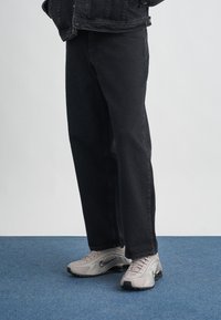 Calças de ganga pretas com um corte reto, acompanhadas de sapatilhas atléticas cinzentas com um padrão texturizado e um acolchoamento proeminente.