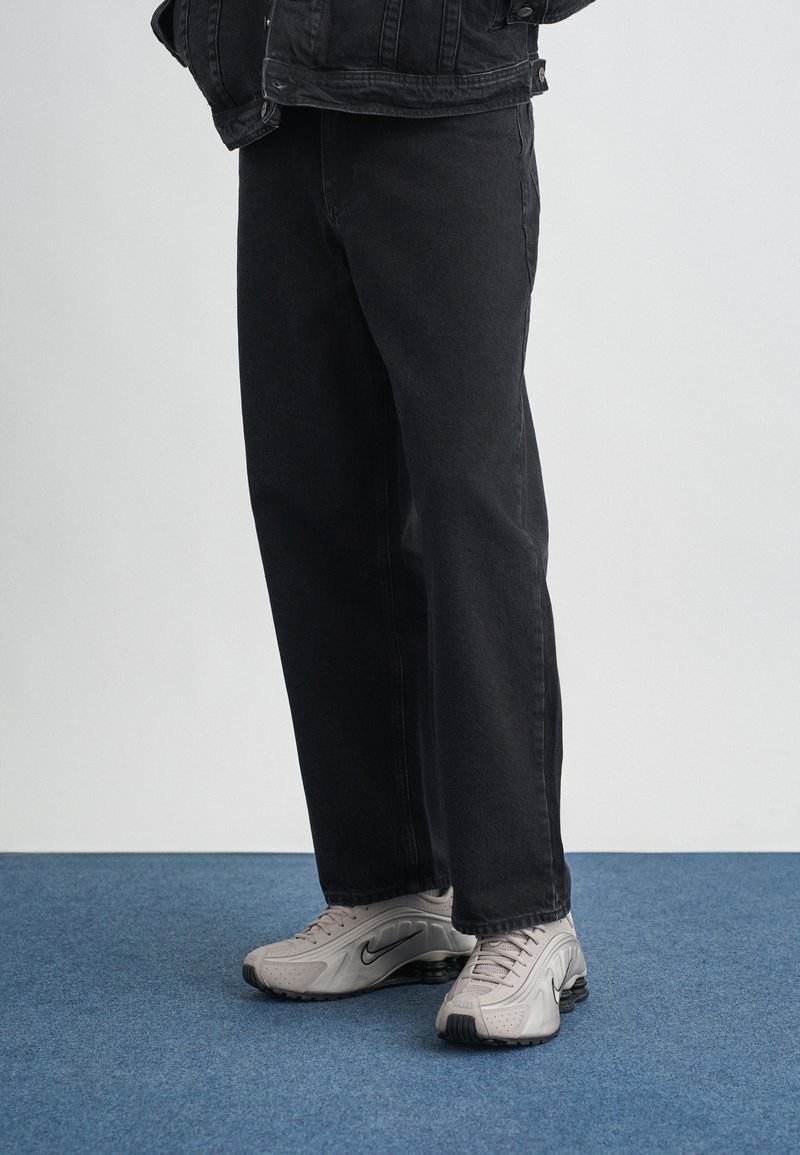 Calças de ganga pretas com um corte reto, acompanhadas de sapatilhas atléticas cinzentas com um padrão texturizado e um acolchoamento proeminente.