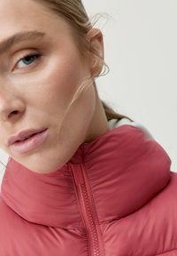 Vadderad jacka med hög krage i en mjuk rosafärg, med en framtida dragkedja och slät текстur. Lätt och varm design.