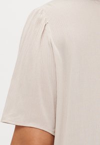 Top beige a maniche corte con una sottile texture stropicciata e un leggero dettaglio a palloncino nella cucitura della spalla.