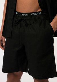 Černé šortky vyrobené z lehké tkaniny s elastickým pasem, na kterém je kontrastní bílé logo "STHUGE". Šňůrka pro nastavení velikosti.