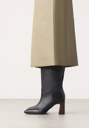 Zwarte leren mid-kuit laarzen met blokhakken, gedragen onder een lange beige rok of jas tegen een effen lichte achtergrond.