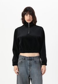 Svart kortermet genser laget av myk velour, med høy krage og halv glidelås, samt elastisk nederkant og mansjetter. Brukt sammen med grå jeans.