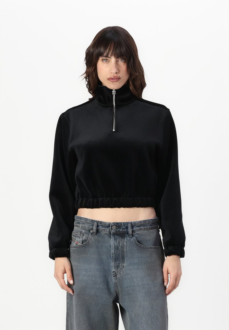 Svart kortermet genser laget av myk velour, med høy krage og halv glidelås, samt elastisk nederkant og mansjetter. Brukt sammen med grå jeans.