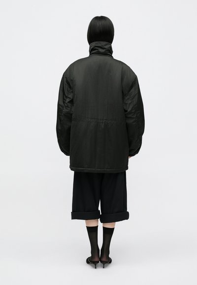 Crna puffer jakna s visokim ovratnikom, širokog kroja, s vidljivim kvačenim detaljima, uparena s skraćenim crnim hlačama i cipelama na petu.