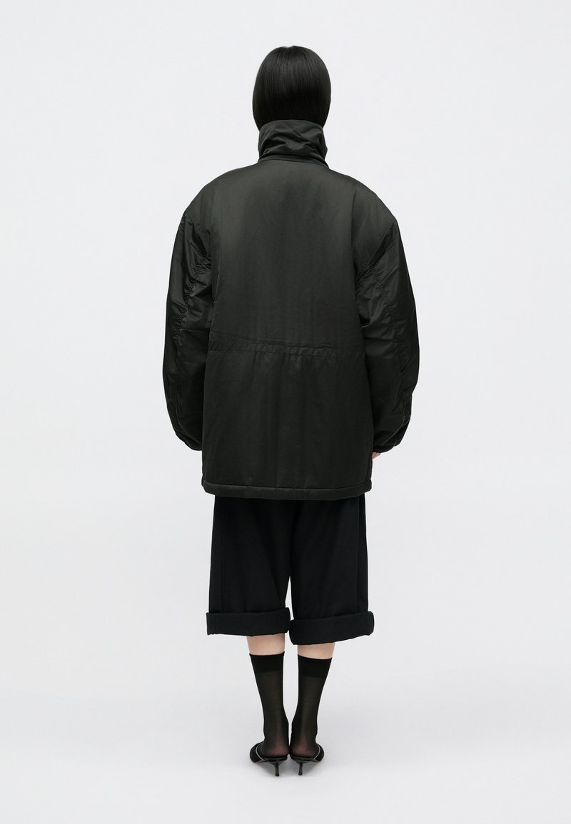 Schwarze Daunenjacke mit hohem Kragen, oversized Schnitt und sichtbar gesteppten Details, kombiniert mit verkürzten schwarzen Hosen und Absatzschuhen.